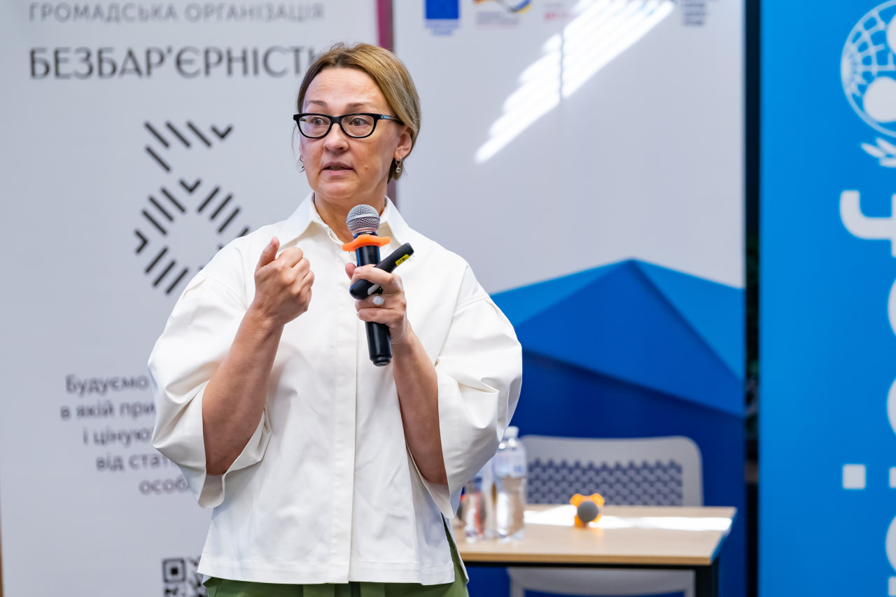 жітка у білій блузці виступає перед аудиторією, в руках тримає мікрофон